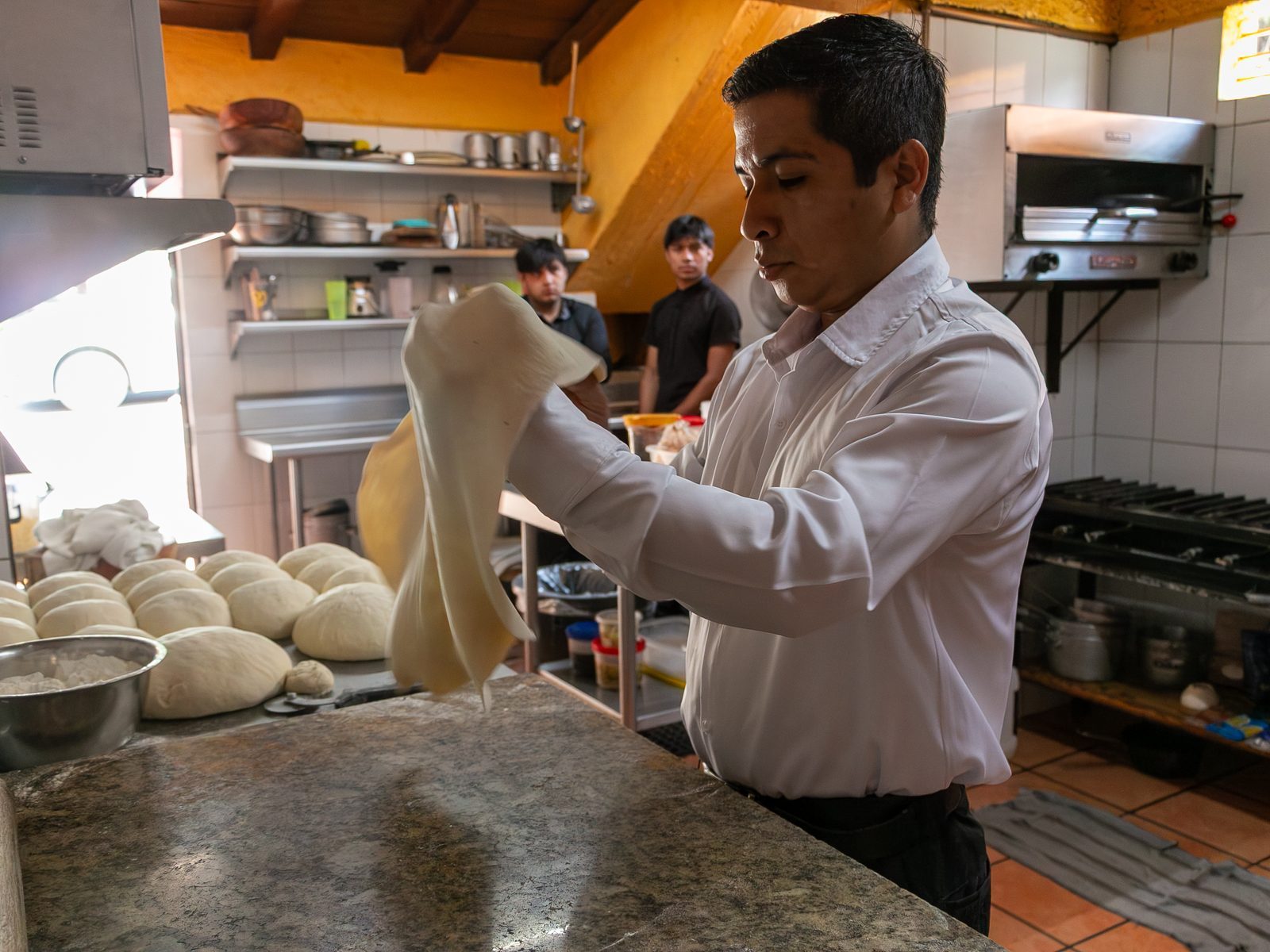 Don Roberto estirando masa de pizza en la cocina, con bolas de masa y el horno de fondo